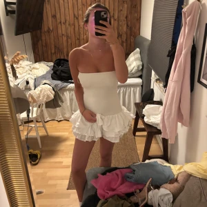Vit axelbandslös volangklänning - Supersöt vit kortklänning med axelbandslös design och flera lager volanger nertill. Tight passform upptill och fluffig kjol som ger en riktigt somrig vibe. Perfekt för dig som vill sticka ut med en lekfull och trendig look.