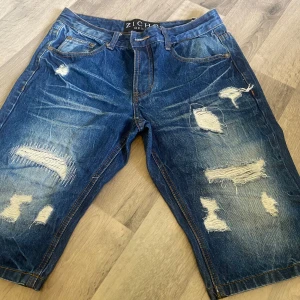 Blå slitna jeansshorts från Zicho Denim - Riktigt snygga blå jeansshorts från Zicho Denim med flera slitningar och trasiga detaljer både fram och bak. Klassisk femficksmodell med normal passform och coola slitna partier som ger en edgy streetkänsla. Perfekta för dig som gillar en avslappnad och trendig stil.