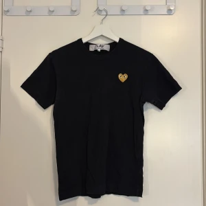 Svart Comme des Garçons Play t-shirt - Svart t-shirt från Comme des Garçons Play med klassiskt guldigt hjärta med ögon broderat på bröstet. T-shirten är i bomull, har rund hals och korta ärmar. Enkel och stilren design som är lätt att matcha.