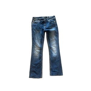 Miss Me blå bootcut jeans med broderi - Snygga blå jeans från Miss Me i bootcut-modell med slitningar och broderade detaljer. Bakfickorna har coola broderier och trasiga partier med camo-tyg under. Tydliga kontrastsömmar och dekorativa knappar ger extra edge. Perfekta för dig som gillar statement-jeans.