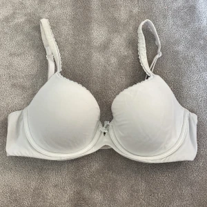 Vit bh från Victoria's Secret - Snygg vit bh från Victoria's Secret i storlek 75C med vadderade kupor och justerbara axelband. Dekorerad med liten spetsrosett och silvrig VS-charm framtill. Bh:n har hakknäppning bak och diskret spetskant längs kuporna. Lite nopprig längst byglarna i bak men går nog lätt att ta bort!