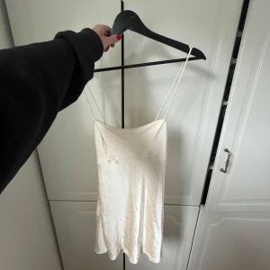 Champagnefärgad satin klänning från Zara - Supersöt kort klänning i champagnefärgad satin från Zara. Den har tunna axelband, snörning i ryggen och en lätt utsvängd passform. Perfekt för dig som gillar minimalistisk och chic stil. Säljes i storlek XS.