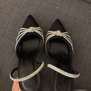 Svarta pumps med strassdetaljer Primark - Snygga svarta pumps från Primark med spetsig tå och hög klack. Skorna har glittrande strassband som bildar en knut framtill och fortsätter runt vristen. Perfekta för att sticka ut med en elegant och trendig look.