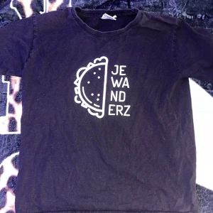 Svart t-shirt med Je Wanderz-tryck - Svart t-shirt med vitt grafiskt tryck framtill som föreställer en taco och texten 'JE WANDERZ'. Klassisk rund halsringning och korta ärmar. Perfekt för dig som gillar unika och roliga prints.