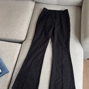 Svarta bootcut kostymbyxor - Snygga svarta kostymbyxor med bootcut passform och pressveck. Byxorna har hällor för bälte och en diskret bakficka. Perfekta för en clean och stilren look. Materialet känns mjukt och följsamt, troligtvis bomull eller polyester.