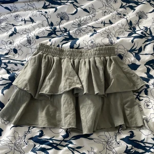 Grön volangkjol från ASOS Design - Supersöt grå kortkjol från ASOS Design med bred resår i midjan och dubbla volanger som ger en riktigt flowy look. Kjolen är gjord i 100% bomull och passar perfekt till perfekt varmt väder. Har dock gått sönder lite, men syns knappt när kjolen är på ❤️