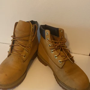 Timberland boots i mocka, beige - Klassiska Timberland boots i beige mocka med robust gummisula och guldfärgade öljetter. Skorna har snörning med tvåfärgade skosnören och vadderad svart kant runt ankeln för extra komfort. Perfekta för dig som gillar streetstyle och outdoor-vibe. Använda 