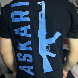 Svart ASKARI t-shirt med blå tryck - Svart t-shirt från ASKARI med stort blått tryck av texten 'ASKARI' och en gevärssiluett på ryggen. Framtill finns samma motiv i mindre format. Klassisk passform och rund hals. Materialet känns som bomull och t-shirten har korta ärmar.