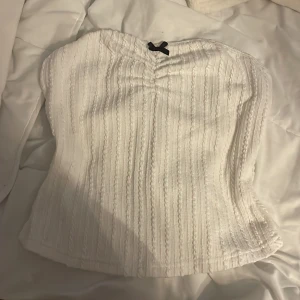 Vit stickad bandeau tubtopp med rosett - Supersöt vit stickad bandeau tubtopp med liten svart rosett framtill. Toppen har ett ribbat mönster och är ärmlös, perfekt för att styla med jeans eller kjol. Mjuk och skön känsla tack vare det stickade materialet.