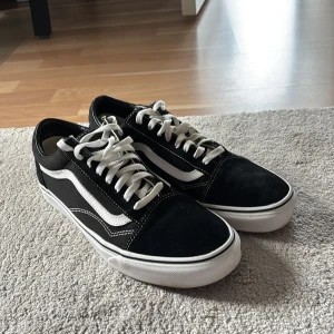 Svarta Vans Old Skool sneakers - Klassiska Vans Old Skool sneakers i svart med vit sula och vit sidosnirk. Skorna har snörning, rund tå och är tillverkade i en mix av mocka och canvas. Perfekta för en streetwear-look och funkar året runt.