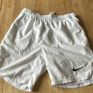 Vita Nike Dri-Fit shorts  - Snygga vita träningsshorts från Nike med Dri-Fit-teknologi. De har elastisk midja med snörning och klassisk svart Nike-logga på benet. Perfekta för sport eller gym och tillverkade i ett lätt och snabbtorkande syntetmaterial.svag fläck som inte går bort!