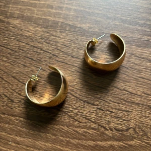 Breda guldiga creoler örhängen - Säljer ett par chunky creoler i guldig metall. De är breda och har en snygg, rundad form som verkligen syns. Perfekta för dig som gillar statement-smycken och vill ha något som drar blickarna till sig. Stängs med klassisk plupp baktill.