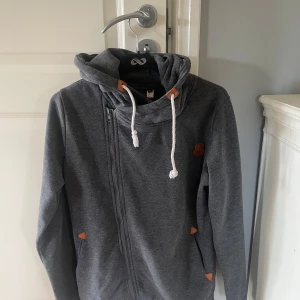 Mörkgrå hoodiejacka från Eltex XL - Mörkgrå hoodiejacka från Eltex i storlek XL. Jackan har dragkedja, snörning i huvan och snygga detaljer i brunt läderimitation. 