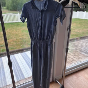 Blå jumpsuit från H&M i bomull - Snygg blå jumpsuit från H&M i mjuk bomull. Den har korta ärmar, krage och knappar framtill. Midjan är markerad med resår för en skön passform. 