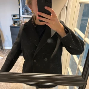 Svart kappa med dubbelknäppning - Stilren svart kappa med klassisk dubbelknäppning och bred krage. Passar både till vintern som vinterkappa men också över söta klänningar/toppar! 