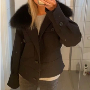 Svart jacka med pälskrage från Meotine - Snygg svart jacka från Meotine med stor fluffig pälskrage. Jackan har två stora fickor framtill, knappar och en boxig, lite kortare passform. Perfekt för dig som vill ha en stilren men ändå statement jacka till höst och vinter.