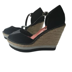 Svarta kilklackar från Vox Shoes - Snygga svarta sandaler från Vox Shoes med hög kilklack i rep och öppen tå. Skorna har en smal rem med spänne runt vristen och ovandel i textil. Perfekta för dig som vill ha en somrig och trendig look med extra höjd.