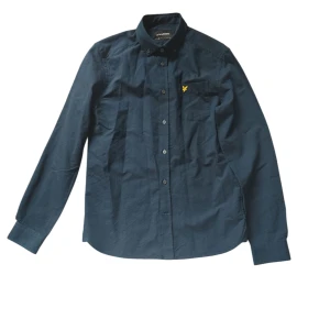 Mörkblå skjorta från Lyle & Scott - Snygg mörkblå skjorta från Lyle & Scott med klassisk button-down krage och gul logga på bröstfickan. Skjortan är i bomull och har långa ärmar samt knappar framtill. Perfekt för dig som gillar stilrena och tidlösa plagg.
