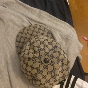 Gucci keps med GG-mönster och röd-blå rand - Snygg keps från Gucci i beige med klassiskt GG-monogram i mörkblått. Kepsen har en röd och marinblå rand på sidan och justerbar rem baktill i svart. Tillverkad i bomullscanvas med svart knapp på toppen. Perfekt för dig som vill sticka ut med lyxig streetstyle.