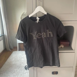 Svart t-shirt med glittertext från H&M - Cool svart t-shirt från H&M med glittrig text 'Yeah' framtill. Klassisk rund halsringning och korta ärmar. Perfekt för dig som vill ha en enkel men ändå statement look. Skön och mjuk bomullskvalitet.
