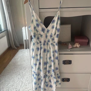 Vit blommig sommarklänning Pull&Bear - Supersöt vit klänning från Pull&Bear i storlek XS med blå och lila blommor. Klänningen har smala axelband, v-ringning och volang nedtill. Den är midilång och har smock i midjan för en skön passform. Perfekt för soliga dagar och chill vibes.