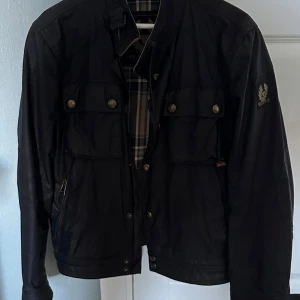 Svart Belstaff jacka med fickor - Snygg svart jacka från Belstaff med klassisk design och flera fickor framtill. Jackan har rutigt innerfoder i blått och grått, hög krage och detaljerade knappar. Perfekt för dig som gillar stilren och tidlös look. 