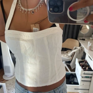 Vit korsett-topp med breda axelband - Säljer en snygg vit korsett-topp med breda axelband och markerade sömmar som ger en cool och strukturerad look. Toppen är croppad och passar perfekt till jeans eller kjol för en trendig outfit. Har en liten fläck.