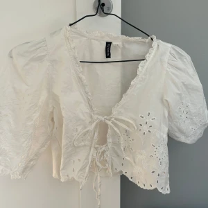 Vit broderad blus från Divided - Superfin vit blus från Divided med broderade blommor och hålbroderier. Modellen har kort puffärm, v-ringning med volangkant och knyt framtill. Perfekt för en romantisk look och passar till både jeans och kjol.