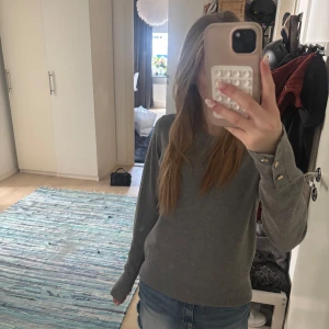 Grå stickad tröja med knappar - Snygg grå stickad tröja med rund halsringning och långa ärmar. Ärmsluten har dekorativa guldfärgade knappar som ger en extra touch. Perfekt till jeans eller kjol för en chill och stilren look.