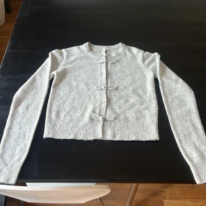 Grå kofta med rosettdetaljer från Nelly - Superfin ljusgrå kofta från Nelly med långa ärmar och snygga rosettdetaljer framtill. Koftan är stickad och har ribbade muddar vid ärmslut och nederkant.