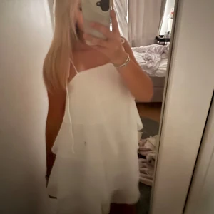 Vit omlottklänning med volanger - Supersöt vit kortklänning med tunna axelband och flera lager volanger som ger en flowy och luftig känsla. Klänningen har omlott-look och är perfekt för varma dagar. Materialet känns lätt och svalt mot huden. Kommer aldrig till användning tyvärr för den är ju så fin. Köptes förra sommaren på Gina för 700 kr. ‼️Det är inte en fläck på klänningen det är spegeln!!