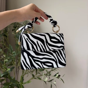 Zebramönstrad handväska med gulddetaljer - Snygg handväska med svartvitt zebramönster och tvinnat handtag med gulddetaljer. Väskan har en stilren, fyrkantig form och stängs med lock. Perfekt för dig som vill sticka ut med en trendig accessoar. OANVÄND
