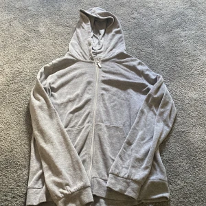 Grå hoodie med dragkedja och huva - Enkel och stilren grå hoodie i storlek 170. Tröjan har hel dragkedja framtill, två stora fickor och en skön huva. Perfekt för chill dagar eller när du vill ha något bekvämt och basic. Tillverkad i mjuk bomullsmix.