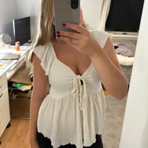 Vit blus med volang och knyt - Supersöt vit blus med djup v-ringning, volangärmar och knyt framtill. Luftig och flowy modell som ger en romantisk vibe. Perfekt till jeans eller kjol för en fräsch look.