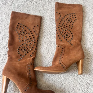 Bruna mockastövlar med nitar och klack - Snygga bruna stövlar i mocka med dekorativa nitar i mönster längs skaftet. Skorna har rund tå och hög klack i trälook, vilket ger en cool och trendig vibe. Perfekta för att lyfta din outfit med lite edge.