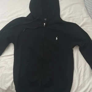 Svart hoodie från Polo Ralph Lauren - Helt ny och oandvänd Svart hoodie från Polo Ralph Lauren säljes pga fel storlek. klassisk vit broderad logga på bröstet. Tröjan har huva med snörning, dragkedja framtill och rymliga fickor. Tillverkad i mjuk bomull, perfekt för chill dagar. Stilren och enkel design som funkar till allt. Nypris 2100