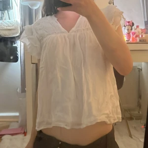 Vit blus med volangärmar - Söt vit blus med v-ringning och volangiga korta ärmar. Lätt och luftig modell med broderade detaljer upptill och lös passform. Perfekt för varma dagar och ger en romantisk vibe.