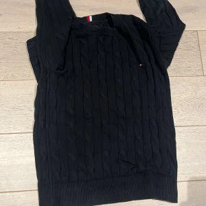 Svart kabelstickad tröja Tommy Hilfiger - Svart kabelstickad tröja från Tommy Hilfiger i klassisk modell med rund hals och ribbade muddar. Liten logga på bröstet och snygg detalj med röd, vit och blå rand i nacken. Perfekt för dig som gillar stilrena och tidlösa plagg.