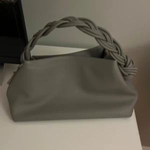 Grå handväska med flätat band - Säljer en stilren grå handväska med ett unikt, tjockt flätat handtag. Väskan har en mjuk, slät yta och är tillverkad i skinnimitation. Perfekt storlek för det viktigaste och ger en cool touch till din outfit. De kommer även med ett axelband som hör till💓har enbart använts en gång så är som ny💗