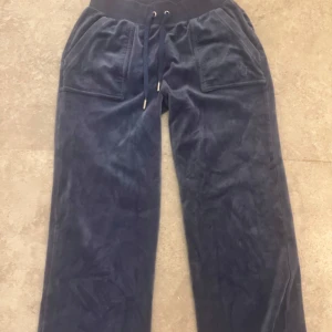 Blå velourbyxor Juicy Couture XS - Mörkblå mjukisbyxor från Juicy Couture i velour med bred resår och snörning i midjan. Byxorna har raka ben, stora fickor fram och diskret broderad logga vid benet. Perfekta för en chill och trendig look.