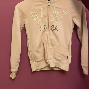 Ljusrosa hoodie från GANT med dragkedja - Säljer en ljusrosa hoodie från GANT med broderad logga och text framtill. Tröjan har huva, långa ärmar och hel dragkedja. Mjuk bomullskänsla och ribbade muddar vid ärmslut och nederkant. Perfekt för en chill och sportig look.