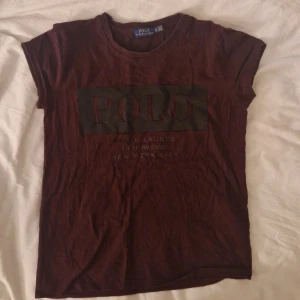 Vinröd t-shirt Polo Ralph Lauren XS - Snygg vinröd t-shirt från Polo Ralph Lauren i storlek XS. T-shirten har korta ärmar och ett stort tryck med 'POLO' på bröstet. Mjuk bomullskvalitet och klassisk rund hals. Perfekt för dig som gillar stilrena märkesplagg.