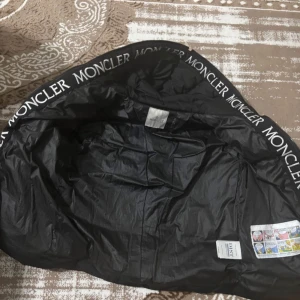 Svart Moncler dunjacka med huva - Snygg svart dunjacka från Moncler med stor huva och tydlig logga längs insidan. Jackan har två sneda dragkedjefickor framtill och klassisk Moncler-patch på ärmen. Perfekt för kalla dagar och riktigt street. Materialet är polyester och dun.
