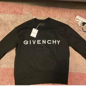 Svart Givenchy sweatshirt med logga - Svart sweatshirt från Givenchy med stor vit logotyp framtill och Givenchy-symbolen broderad på ryggen. Tröjan har rund hals, långa ärmar och ribbade muddar. Perfekt för dig som gillar exklusiva streetwear-vibbar och vill sticka ut med designerplagg.