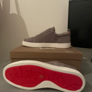 Grå Louboutin - Snygga sneakers från Christian Louboutin i grå mocka med klassisk röd sula och vita kanter. Skorna har rund tå, snörning och diskreta detaljer. Perfekta för dig som vill ha en stilren och lyxig look med en touch av streetstyle.