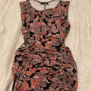 Mönstrad kort klänning från Bershka - Säljer en färgstark kort klänning från Bershka med paisley-inspirerat mönster i svart, rött, rosa och beige. Klänningen är ärmlös, har rund hals och snygga rynkade detaljer längs sidorna för en cool siluett. Perfekt för dig som gillar att sticka ut med din stil.