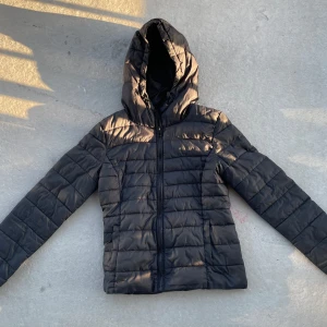 Svart pufferjacka med huva från Only - Snygg svart pufferjacka med huva och dragkedja framtill. Jackan har quiltad design och raka linjer, perfekt för kyliga dagar. Långärmad modell med praktiska fickor på sidorna. Enkel och stilren look som funkar till det mesta.