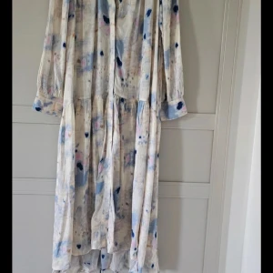 Långklänning med blå och rosa mönster - Säljer en flowy långklänning med ljus bas och abstrakt mönster i blått, rosa och beige. Klänningen har långa ärmar med knappslut, v-ringad hals och volangdetaljer vid midjan. Materialet känns lätt och luftigt, perfekt för varmare dagar.