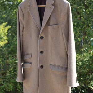 Beige ullkavaj med knappar - Klassisk grå rock i ull med fyra fickor och snygga knappar fram samt på ärmsluten. Fodret är mörklila och materialet känns mjukt och lyxigt. Passformen är normal och rocken har en stilren krage med spetsiga slag.