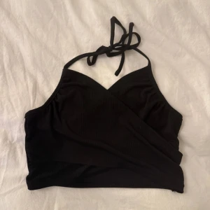 Svart omlott halterneck topp - Svart croppad halterneck topp med omlott-design och knyt i nacken. Snygg och enkel modell som passar perfekt till höga jeans eller kjol. Ribbad struktur ger en extra touch.